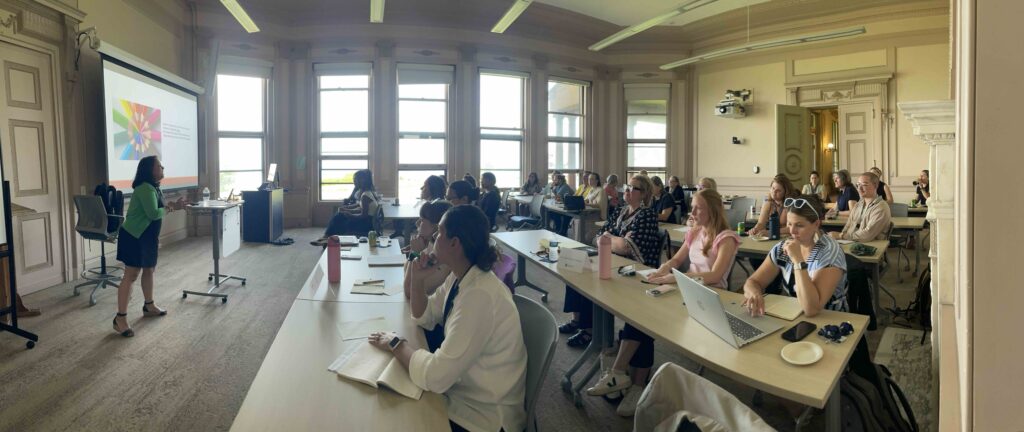 Workshop attendees listen to a presentation during the RIRAC Research Learning Community Series at Salve Regina University. A speaker presents at the front of the room beside a projected slide while participants take notes and follow along from classroom-style tables.