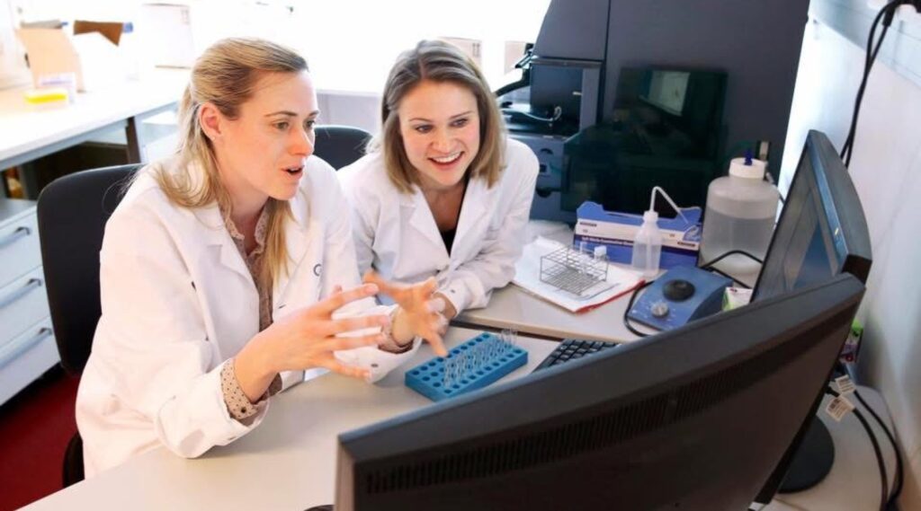 Jillian Wise and another collegue sitting in white lab coats discussing something they are viewing on the computer in front of them.
