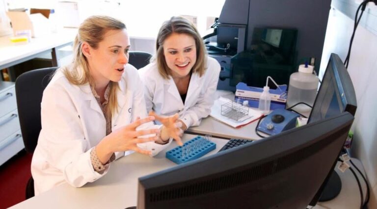 Jillian Wise and another collegue sitting in white lab coats discussing something they are viewing on the computer in front of them.
