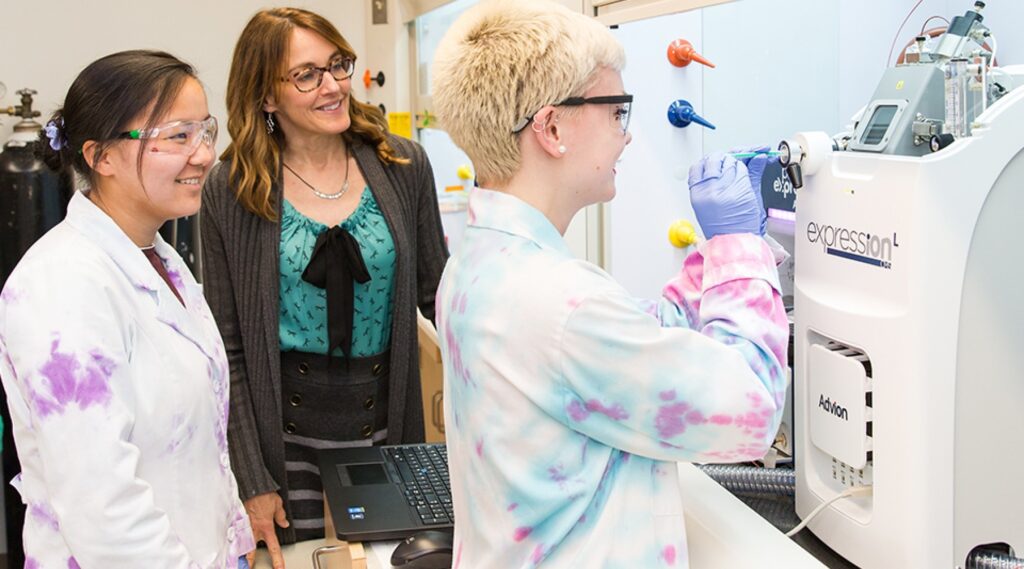 Two students wearing safety glasses and laboratory coats work with a scientific instrument in a research laboratory while an instructor observes. One student operates the equipment as a laptop sits nearby, demonstrating hands-on chemistry research and mentorship in a STEM learning environment.