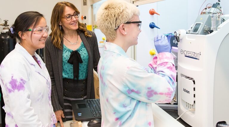 Two students wearing safety glasses and laboratory coats work with a scientific instrument in a research laboratory while an instructor observes. One student operates the equipment as a laptop sits nearby, demonstrating hands-on chemistry research and mentorship in a STEM learning environment.